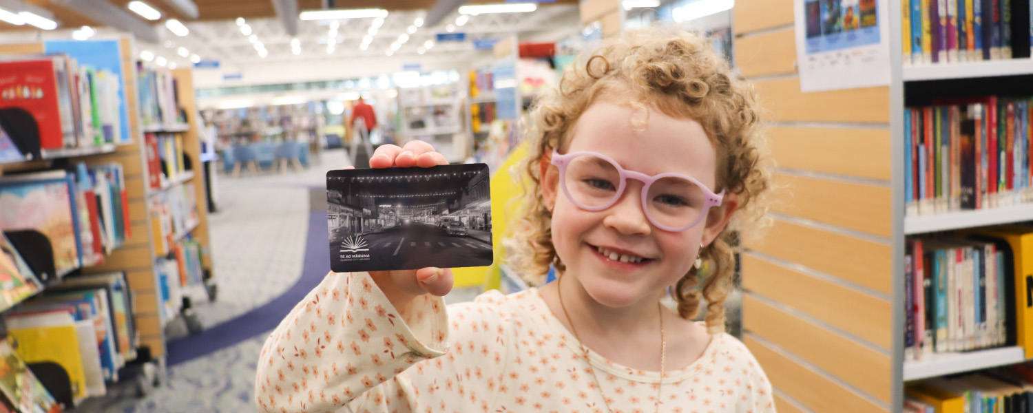 A Tauranga library card held up by a happy, smiling child