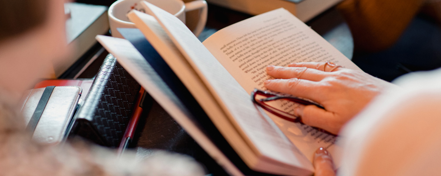 Close up of a book open with a hand on the page holding a pair of reading glasses