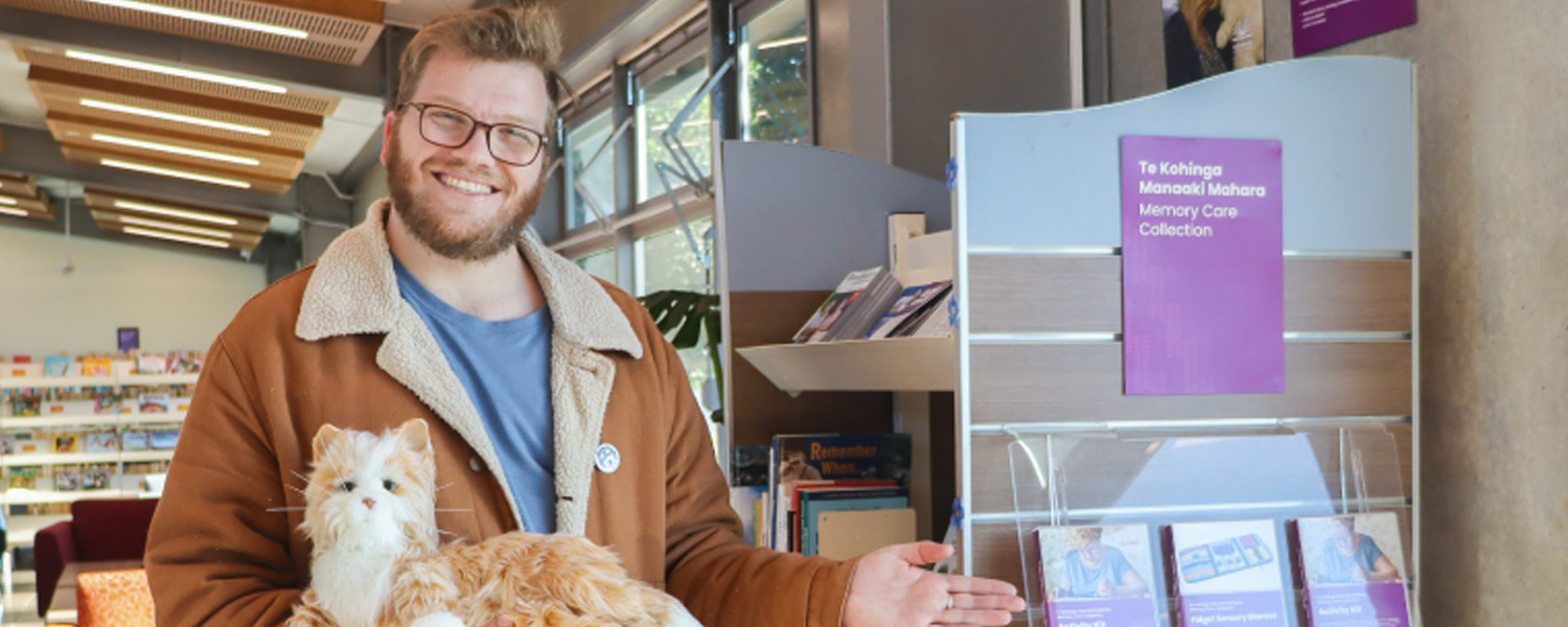 A smiling man gesturing towards a book stand with purple memory care resources